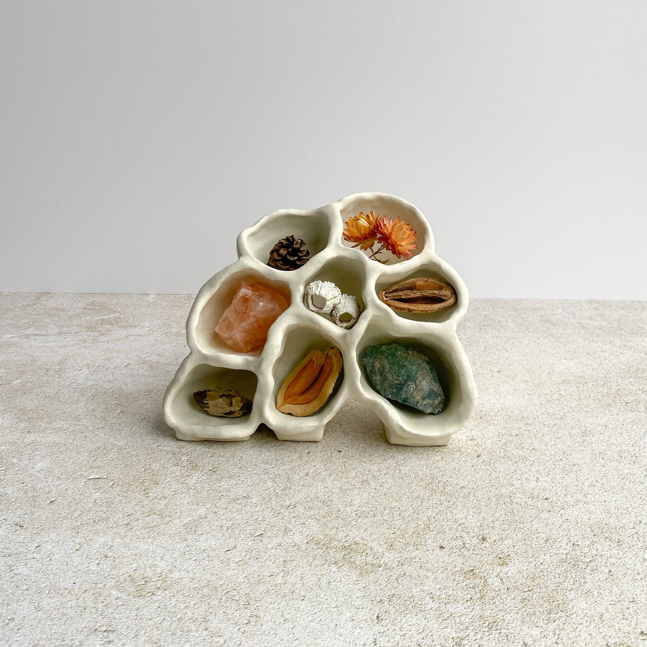 White ceramic sculpture vessel with compartments filled with various natural items on a light surface.