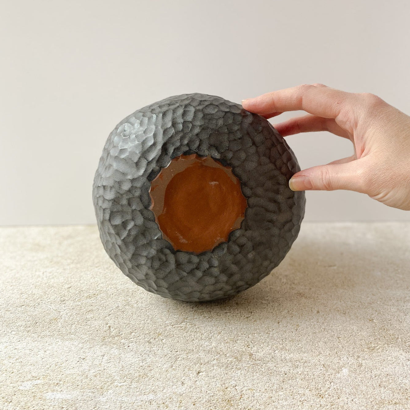 Hand displaying the bottom of a textured ceramic vase on a light surface with a neutral background