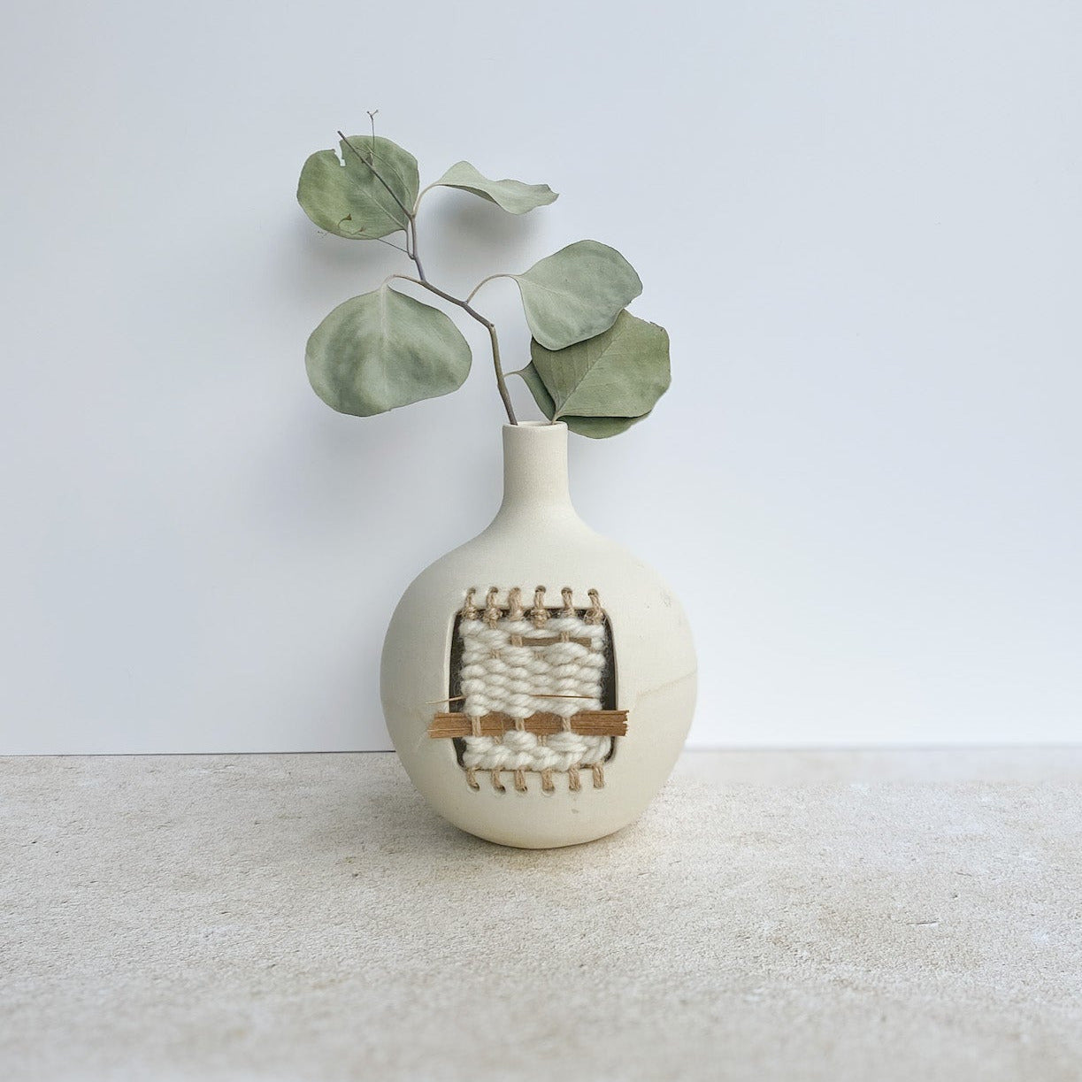 White vase with woven design and eucalyptus leaves on a white surface