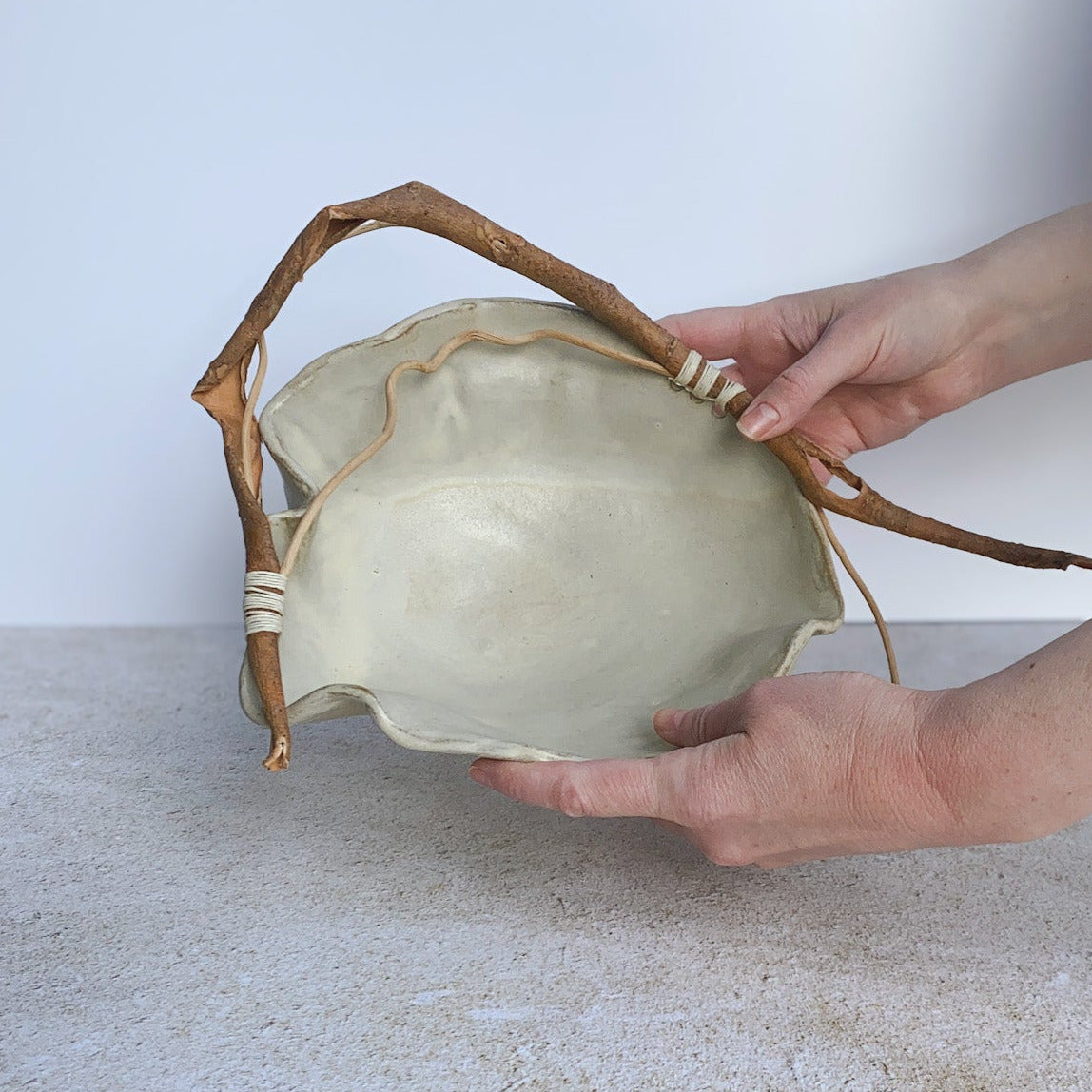 Hand holding a light-colored ceramic bowl with natural fiber handle on a textured surface.
