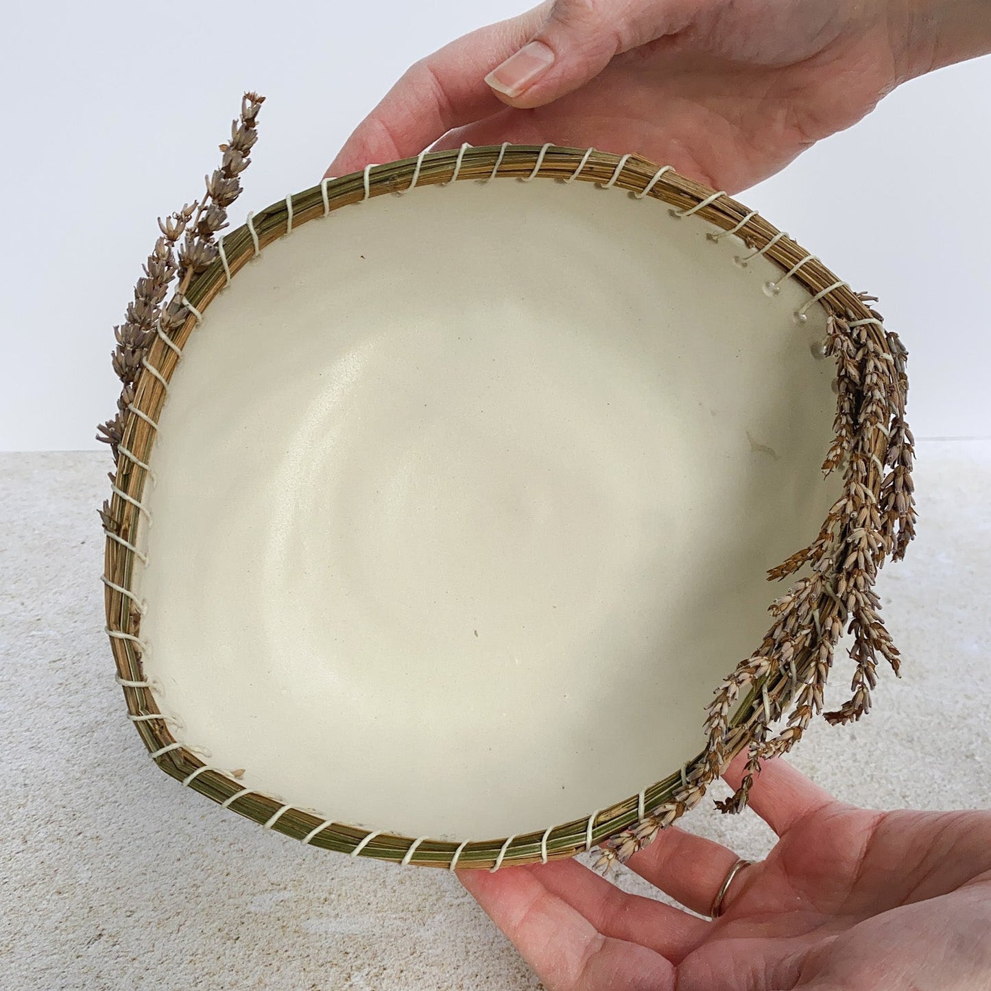 Hand holding a white ceramic bowl with a woven lavender rim on a textured surface