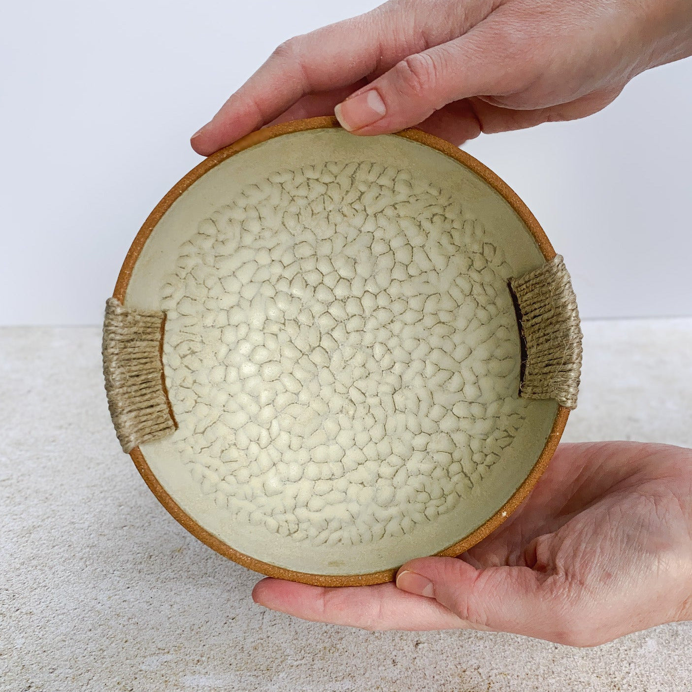 Hand holding a textured ceramic bowl with woven handles on a light surface.