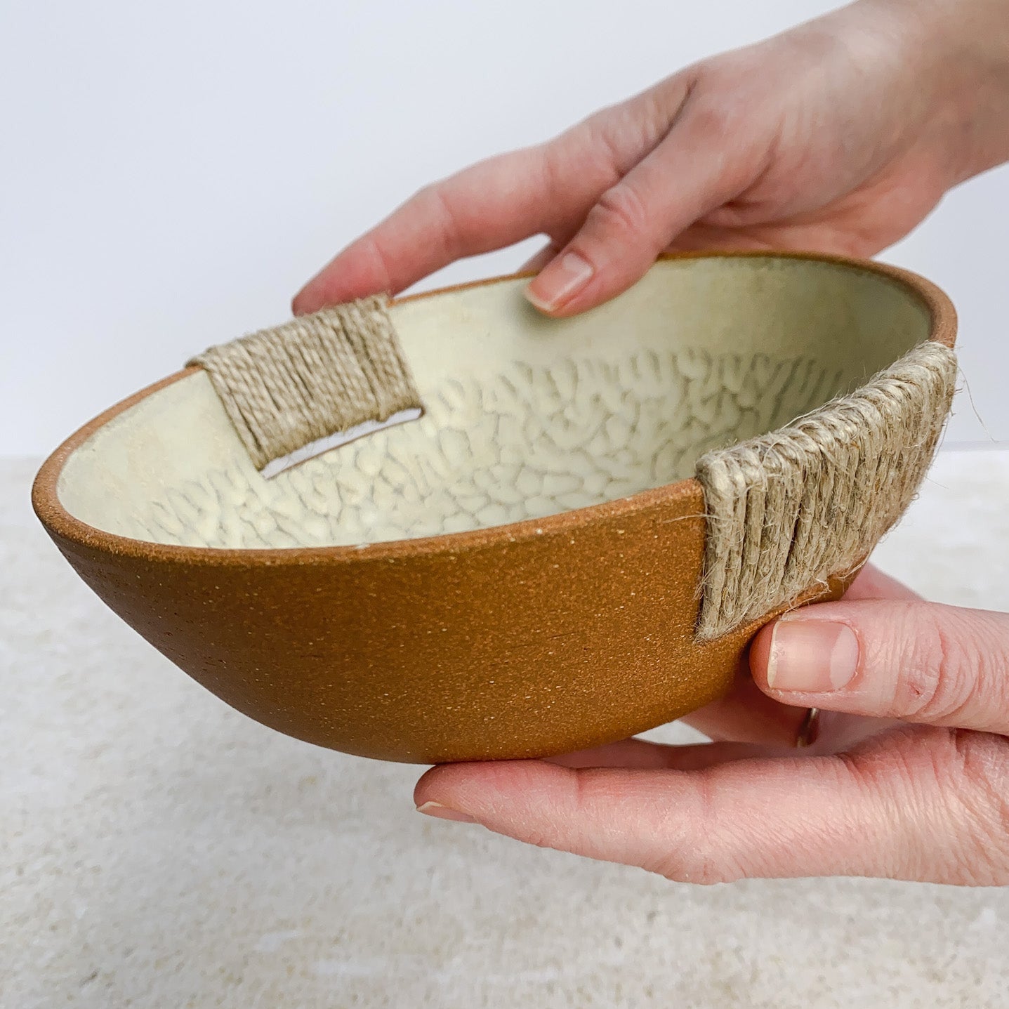 Ceramic bowl with textured interior and natural fiber exterior held by a hand on a light background