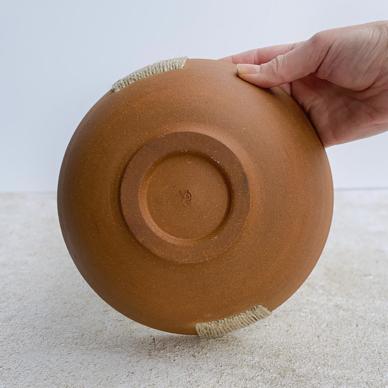 Hand holding an unglazed ceramic bowl on a textured surface with a light background