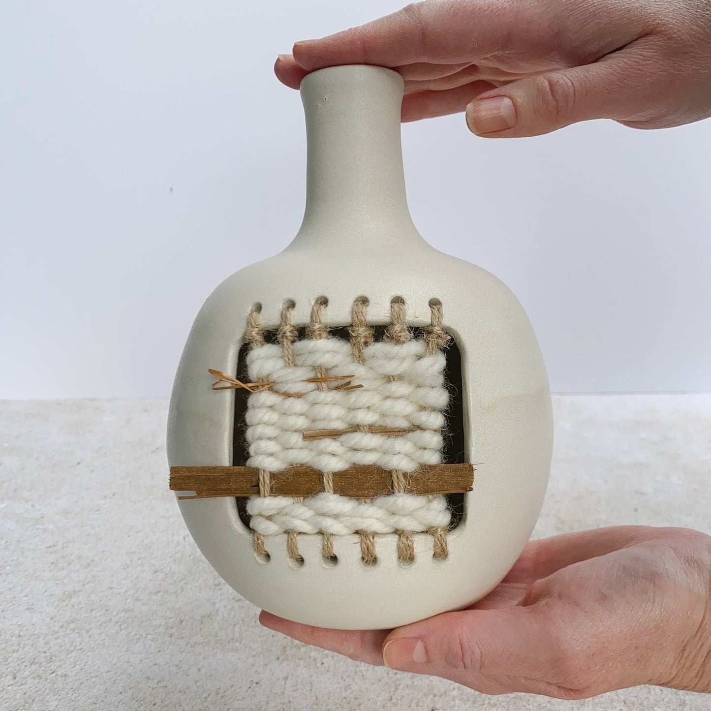 Hands holding a ceramic vase with woven design against a plain background