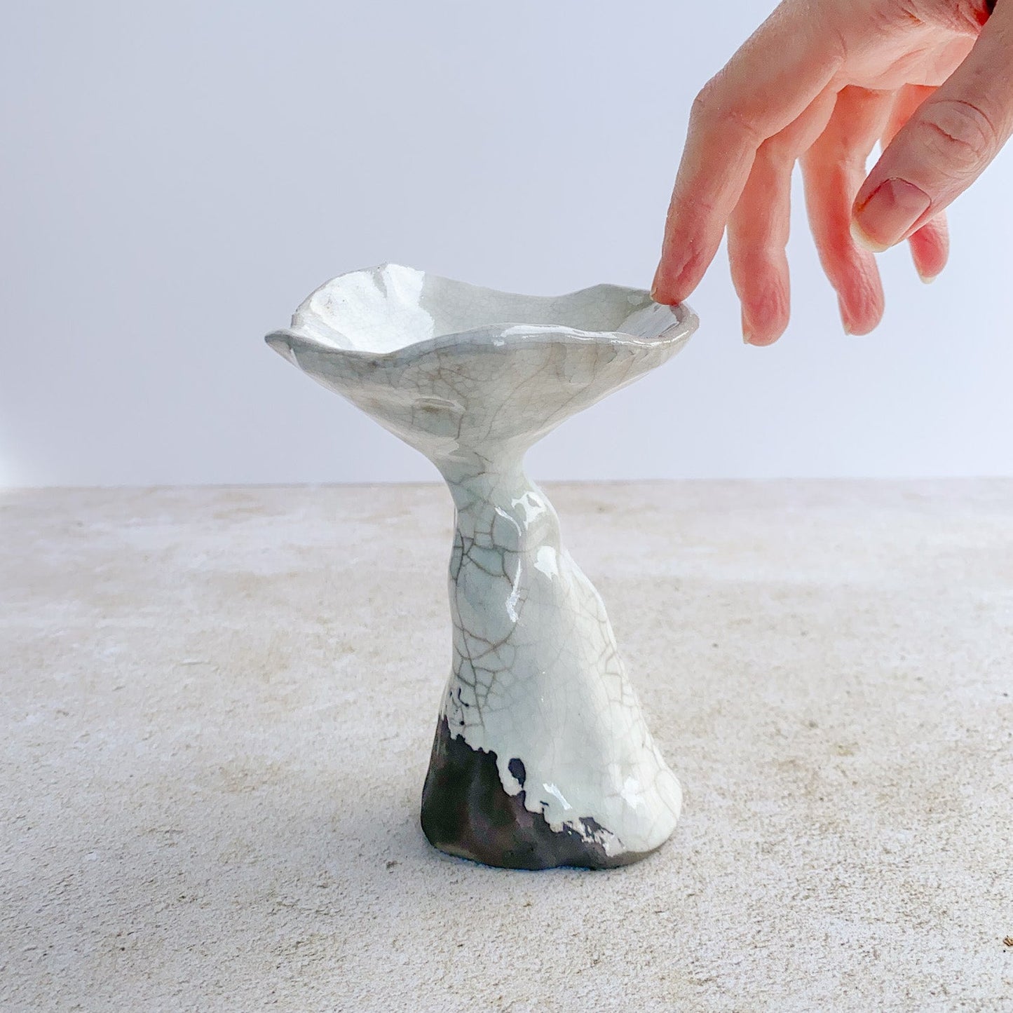Hand reaching toward a ceramic mushroom sculpture on a textured surface on a light background