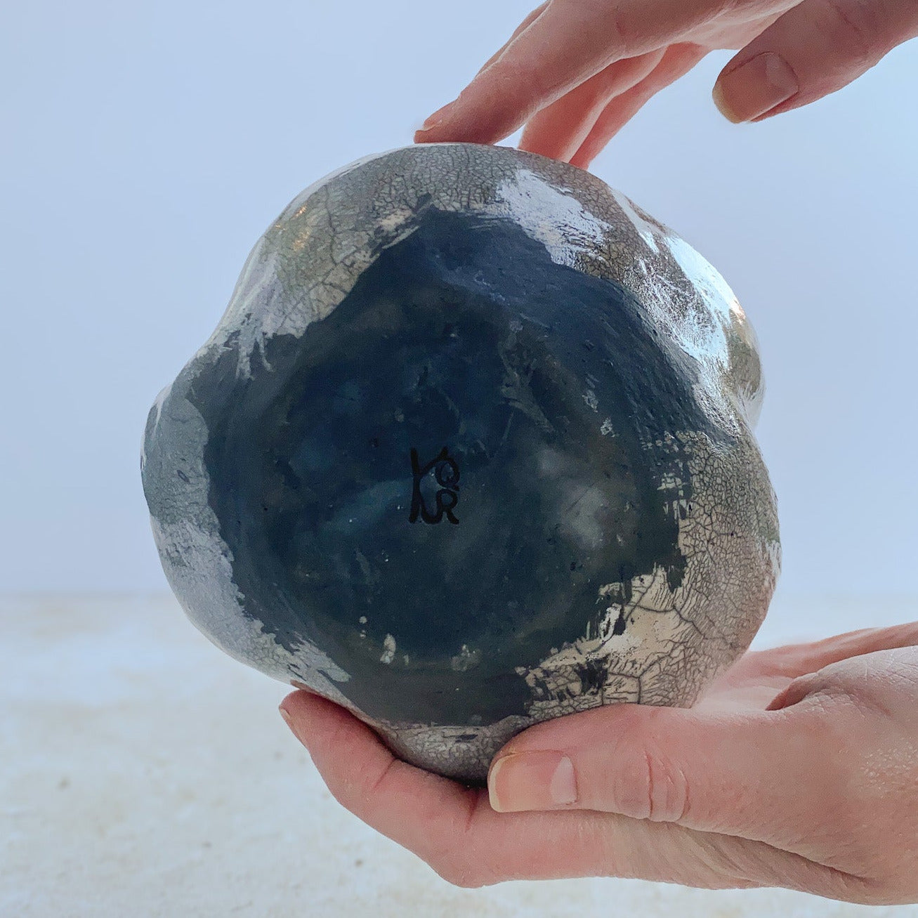 Hands displaying the bottom of a raku ceramic vase on a light background.