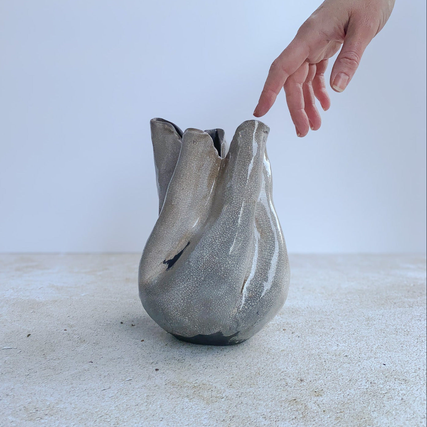Handmade ceramic vase on a white surface with a hand reaching towards it.