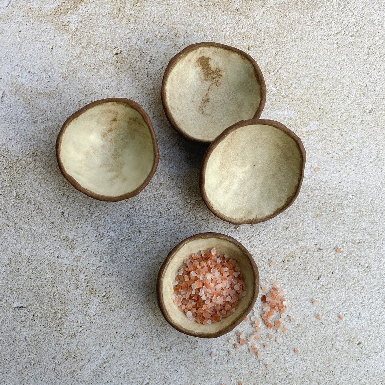 Three small ceramic bowls on a textured surface with pink salt inside one of them.