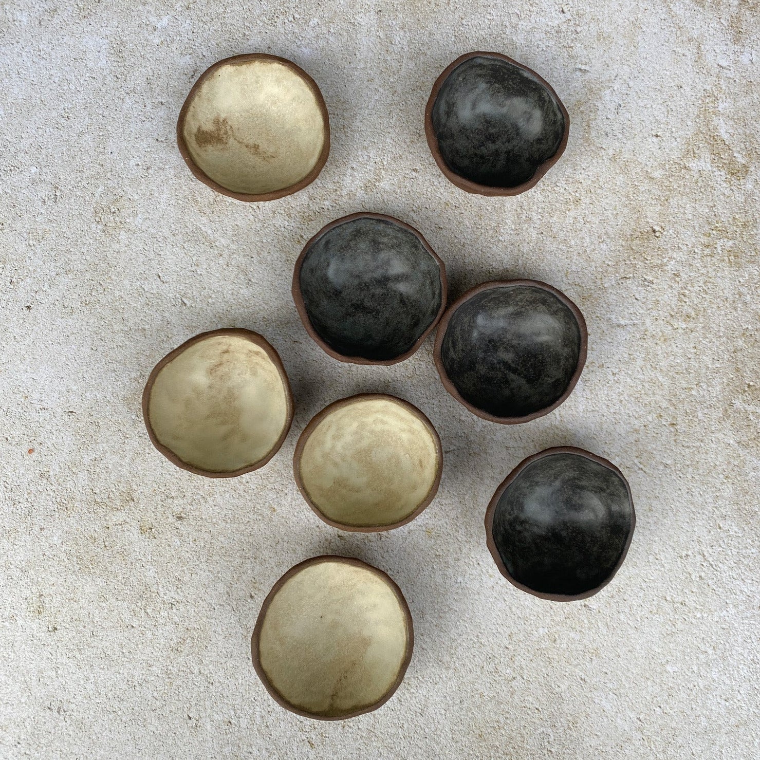 Small ceramic bowls, alternating between black and off white, on a textured stone surface.