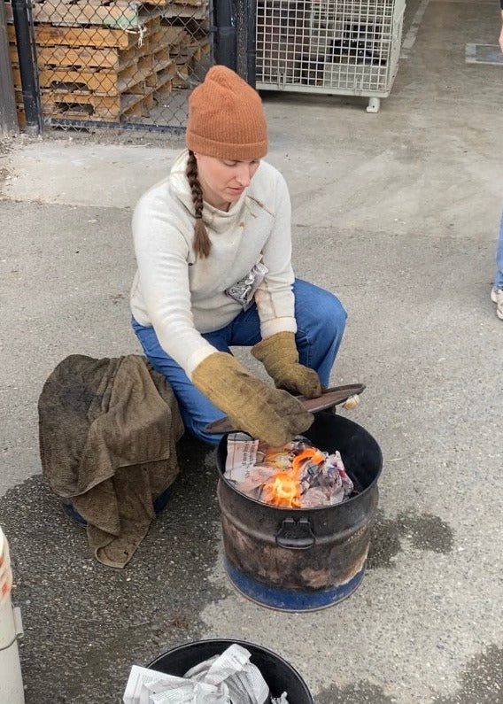 Person sitting on cement using fire for raku pottery in a black container outdoors.