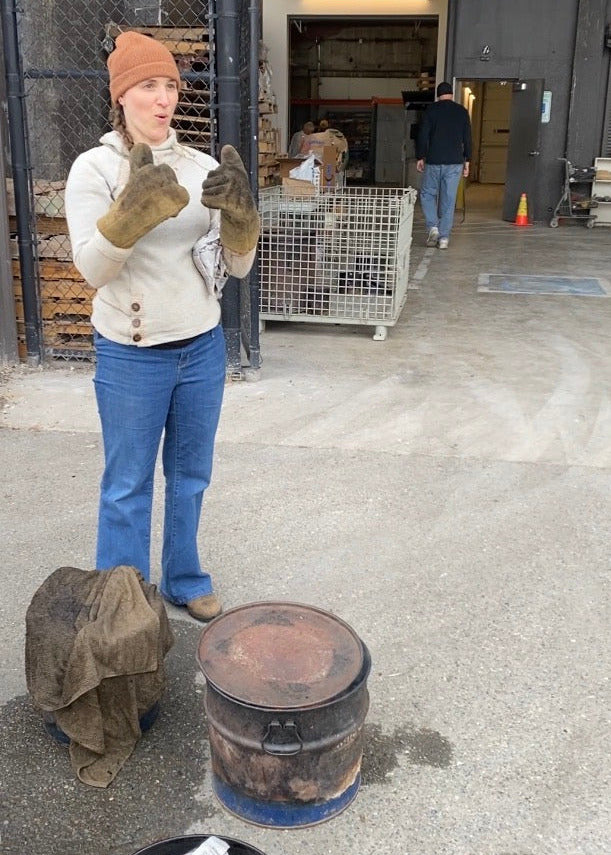 Person in a warehouse setting wearing gloves and a hat, with a barrel and other items on the ground.