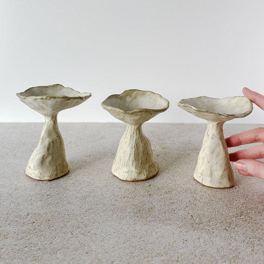 Three ceramic mushroom ring dishes on a neutral surface with a hand reaching towards one of them.